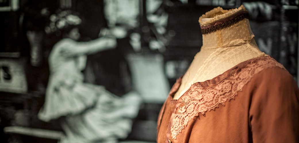 Verschiedene Kleider im Sans-Ventre-Stil auf Schneiderpuppen drapiert. Dahinter ist ein Hemd mit einer Weste und einem Strohhut zu sehen, welche sich ebenfalls auf einer Schneiderpuppe befinden.
