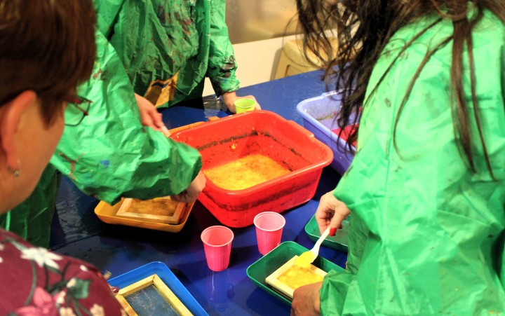 Zwei Personen in grünen Schutzkitteln schöpfen an einem Tisch handgemachtes Papier aus einer geblichen Wasser-Mischung. Vor ihnen stehen Schöpfrahmen, Kunststoffwanne und Becher. Neben den beiden Personen steht eine dritte Person, die den beiden zuguckt.
