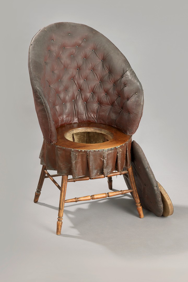 Foto zeigt einen gepolsterten braunen Stuhl mit offenem Sitz in dem sich das Plumpsklo befindet