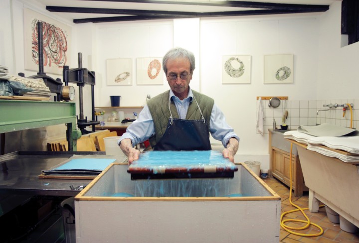 John Gerard steht an einer Schöpfbütte und hält ein rechteckiges Sieb mit blauem Papierbrei in einem Atelier mit Kunstwerken an der Wand.