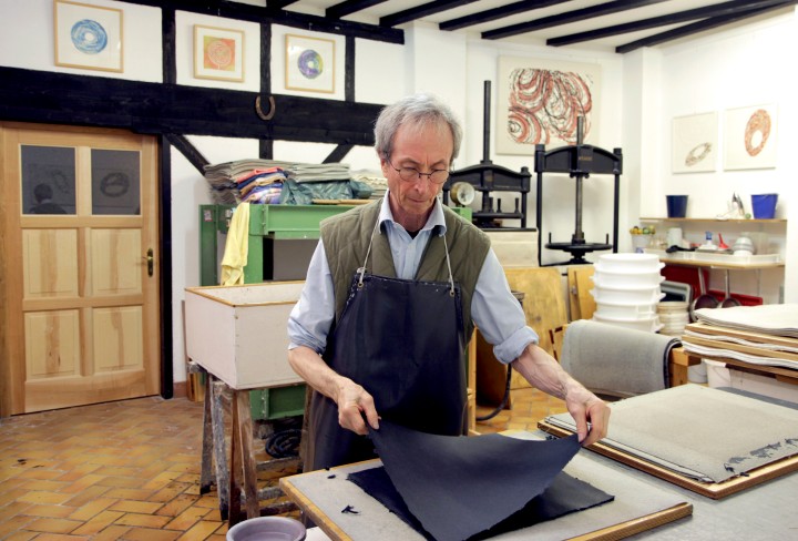 John Gerard mit Schürze faltet schwarzes Papier in einem Atelier mit Holzdecke und gerahmten Bildern an den Wänden.
