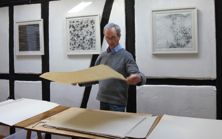 John Gerard steht vor einem flachen Wasserbecken und hält ein rechteckiges Sieb mit blauem Papierbrei in einer Werkstatt mit Kunstwerken an der Wand.