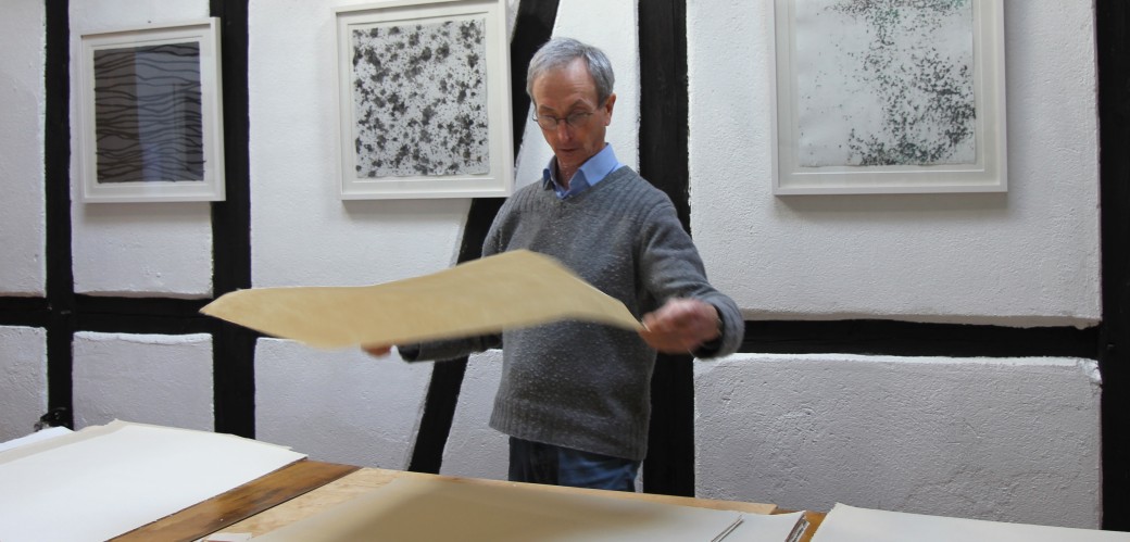 Der Künstler John Gerard sortiert große helle Papierbögen auf einem Tisch.