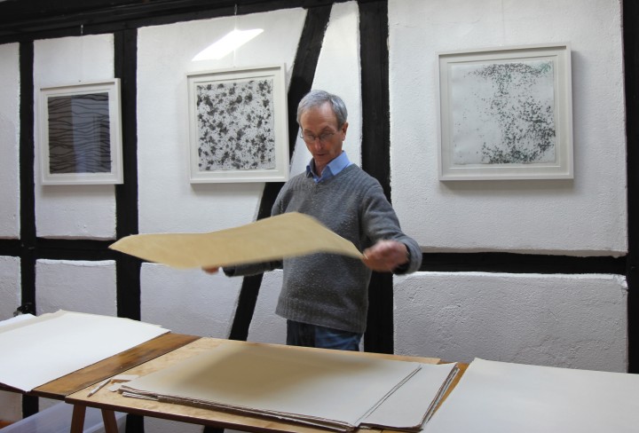 John Gerard steht vor einem flachen Wasserbecken und hält ein rechteckiges Sieb mit blauem Papierbrei in einer Werkstatt mit Kunstwerken an der Wand.