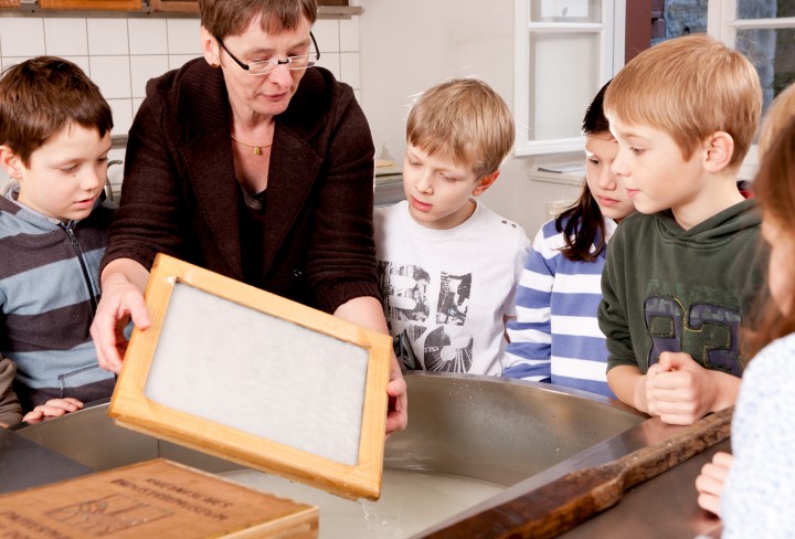 Eine Mitarbeiterin des Museums zeigt einer Gruppe Kinder, wie man Papier schöpft.