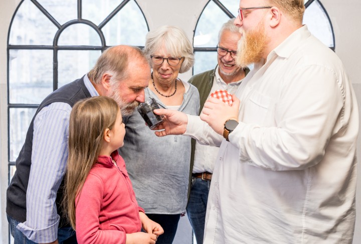 Ein Mann hält einem älterem Herrn ein Glas mit etwas zum Riechen unter die Nase. Ein Mädchen, eine ältere Frau und ein anderer älterer Mann gucken dabei zu.