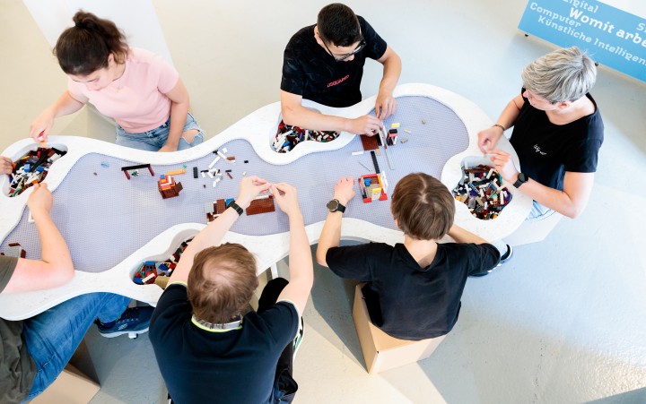 Eine Gruppe junger Menschen steht um einen organisch geformten Basteltisch und baut mit Lego.