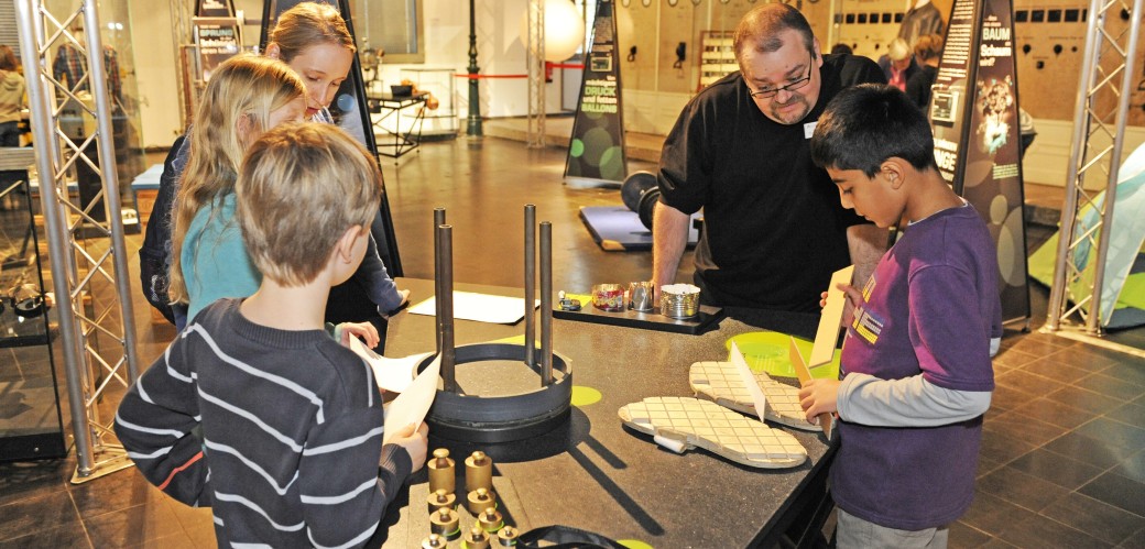 Eine Gruppe Kinder experimentiert an einer Station. Ein Museumsmitarbeiter hilft ihnen.