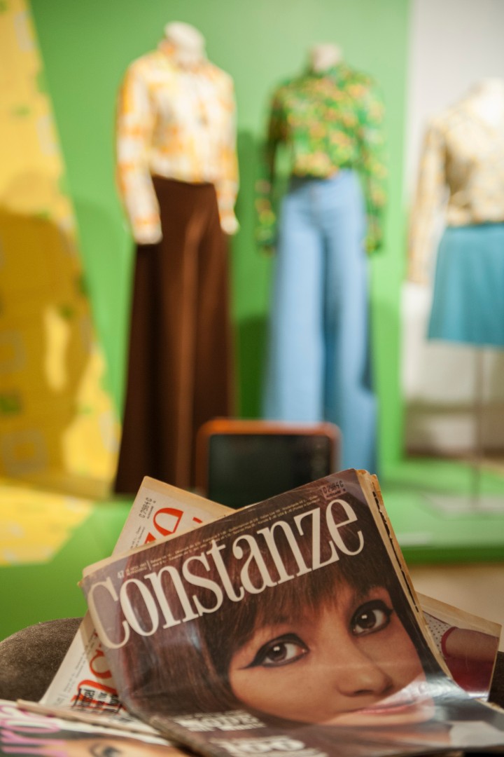 Im Vordergrund liegt eine Modezeitschrift und im Hintergrund stehen Hosen mit Blusen auf Figurinen