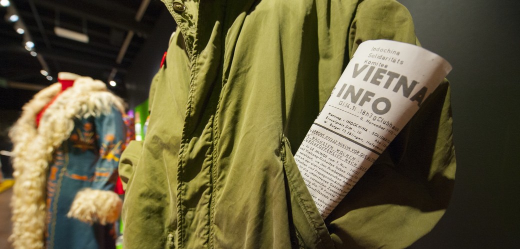 Ein grüner Parka, in dessen Jackentasche eine Zeitung mit der Überschrift "Vietnam Info" eingerollt ist.