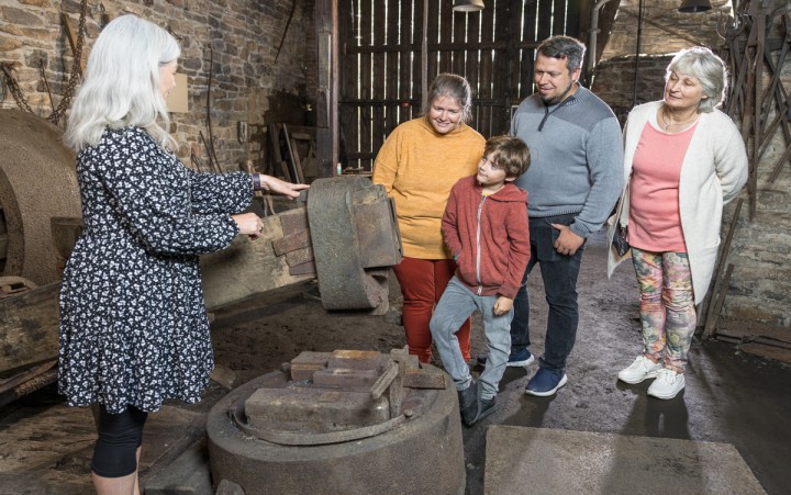 Frau mit grauen Haaren erklärt vier Personen in einer alten Schmiede eine große Maschine.