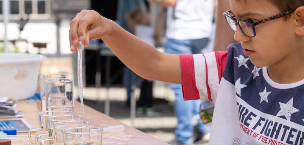 Ein Junge mit Brille träufelt mit einer Pipette Wasser in sechs nebeneinander aufgereihte Gläser.