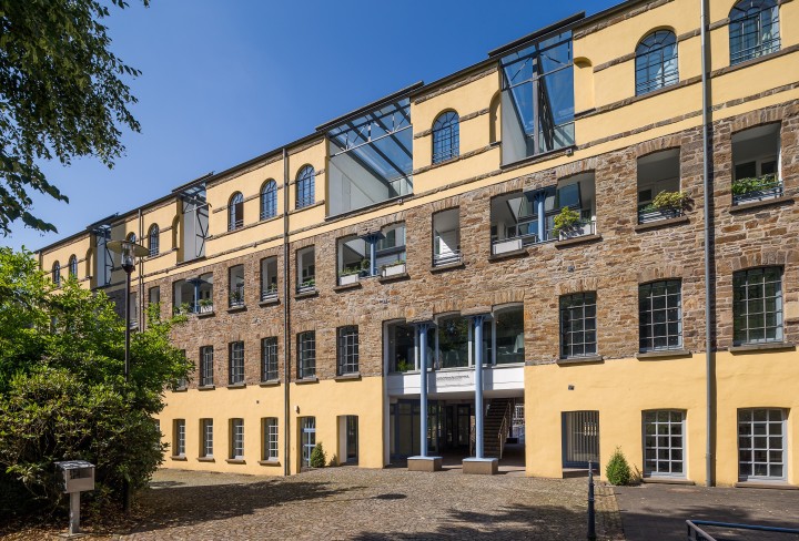 Foto zeigt das gelbe vierstöckige Fabrikgebäude mit grauem Hof davor