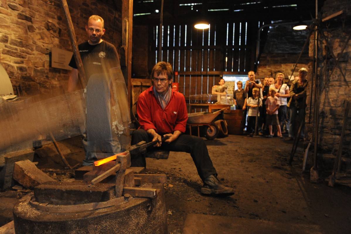 Museum blacksmith and helpers at work on the forge hammer, group of children is watching.
