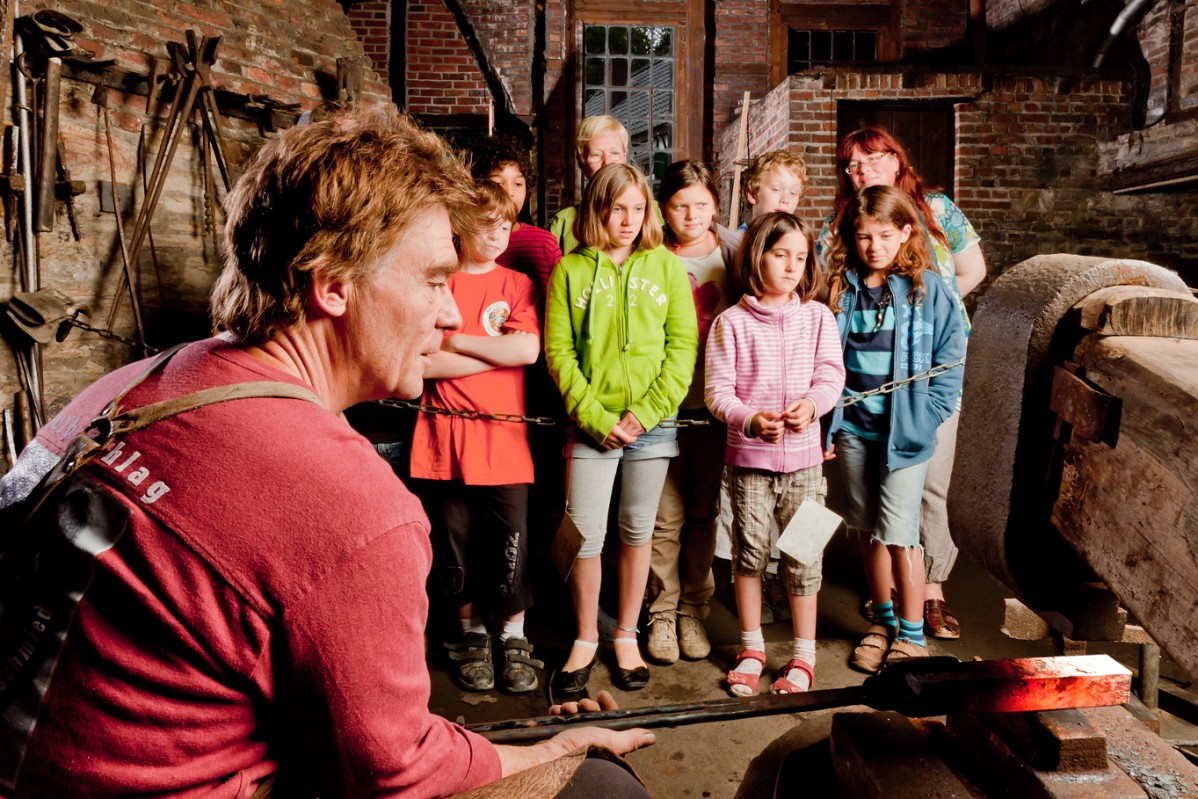 A group of children watches the museum blacksmith forging a workpiece.