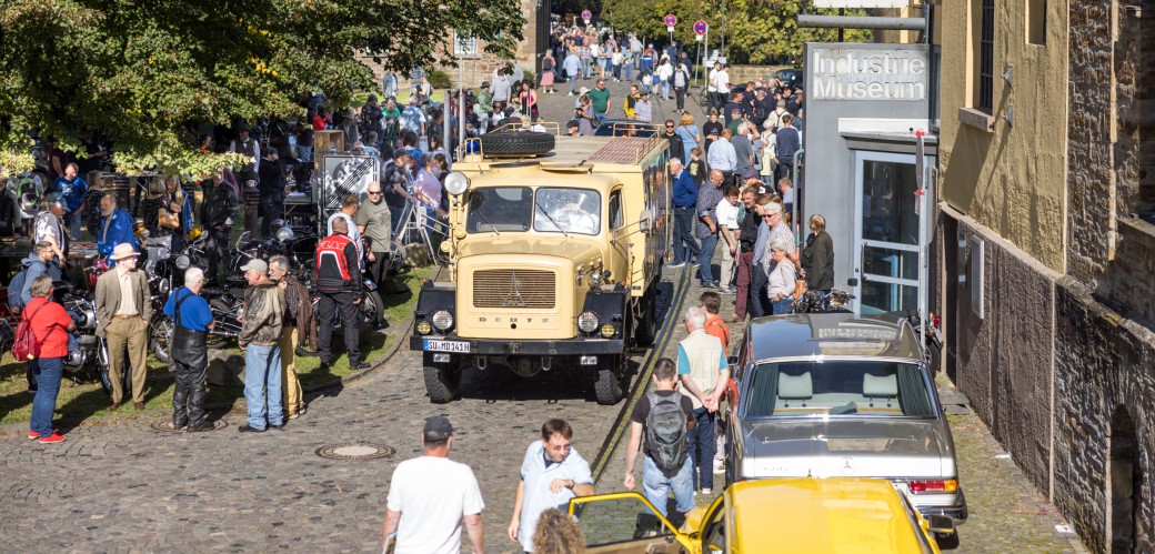 Eine große Gruppe von Menschen versammelt sich auf dem Außengelände des LVR-Industriemuseums in Engelskirchen. Auf dem Weg sieht man einen historischen LKW. Am Wegerand, vor dem Museumseingang, parken zwei historische PKW. 