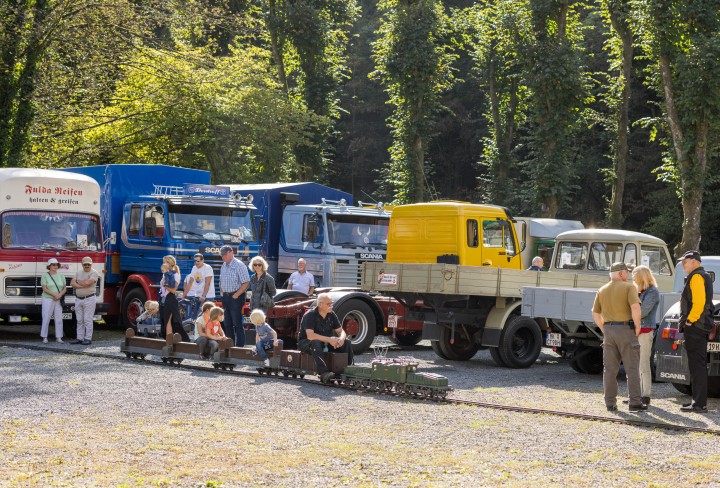 Mehrere historische LKW auf einem Platz, davor Besuchende und eine Kinder-Dampfeisenbahn