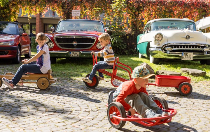 Kinder auf verschiedenen Kinder-Fahrzeugen vor historischen Autos
