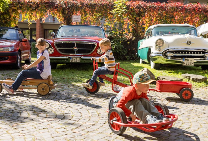 Drei Kinder auf Dreirädern vor herbstlicher Kulisse mit Oldtimer-PKW
