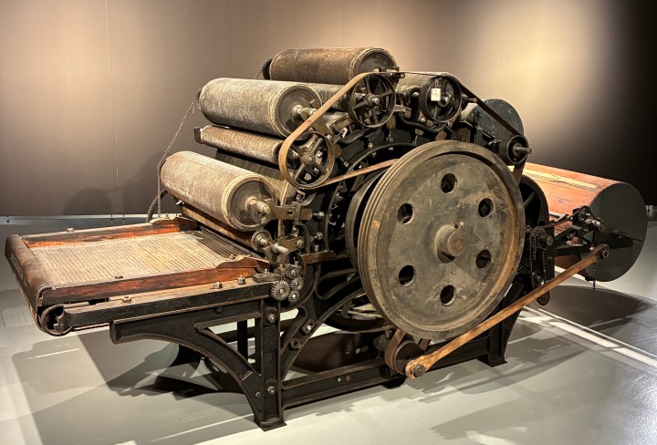 Große historische Textilmaschine aus Metall und Holz, ein sogenannter Krempel. Die Maschine steht frei im Raum eines Museums. Mehrere übereinander angeordnete Walzen sind oben auf der Maschine. Vorne befindet sich ein großes, rundes Schwungrad mit Löchern, das über Lederriemen und Zahnräder mit den Walzen verbunden ist. Unten trägt ein stabiles schwarzes Metallgestell die Konstruktion. Links ist eine flache Zuführfläche mit einem hölzernen Rahmen zu sehen, über die vermutlich Rohfasern in die Maschine eingezogen wurden. 