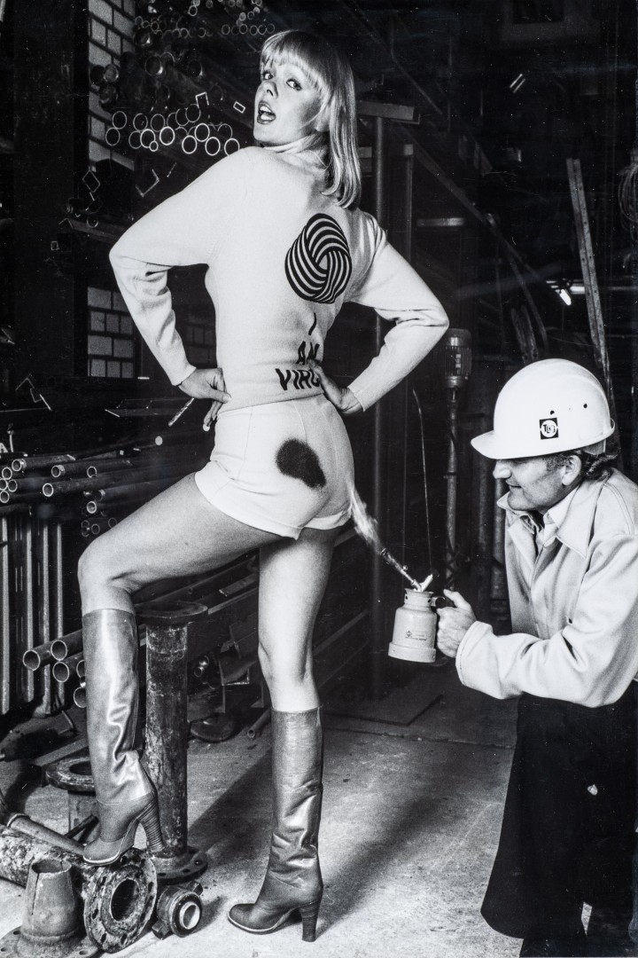 Schwarz-weißes Foto: Eine Frau in kurzen Shorts, langärmligem Oberteil und kniehohen Stiefeln steht in einer Werkhalle auf einem Metallteil und blickt über die Schulter zur Kamera. Auf ihrem Rücken ist ein großes Wollknäuel-Logo zu sehen. Ein Mann mit Schutzhelm kniet hinter ihr und richtet ein Gerät mit Flamme auf die Rückseite ihrer Shorts, wodurch ein dunkler Fleck entsteht. Im Hintergrund liegen Rohre und Metallteile, die die industrielle Umgebung betonen.