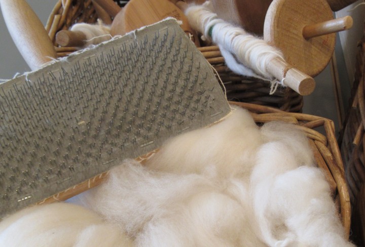 white wool lies in a bast basket. Next to it is a carding brush with lots of small hooks and empty wooden thread spools