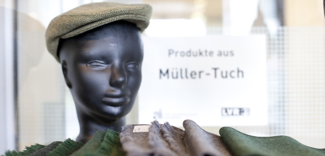 Im Vordergrund liegen flach gewebte Schals in grünen und grauen Farben. Ein schwarzer Modellkopf trägt eine beige Schiebermütze. Im Hintergrund hängt ein Schild "Produkte aus Müller-Tuch".