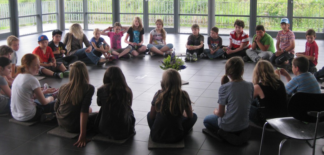 Many children are sitting in a circle on the floor of a large room