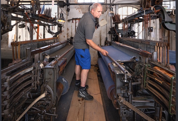 Mann in einem grauem T-Shirt und blauer Shorts bedient eine Webmaschine in der Tuchfabrik Müller.