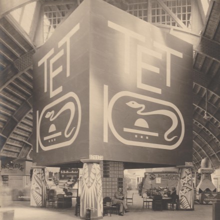 Schwarz-weiß Foto einer Halle. In der Mitte ist ein würfelförmiger Aufbau, auf dem "TET" geschrieben steht und ein Logo einer Schlange zu sehen ist. Unter dem Würfel sitzen einige Personen an Tischen.