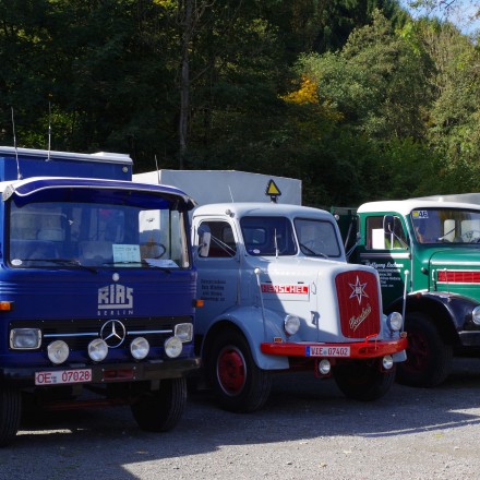 Drei historische LKW stehen nebeneinander auf einem Parkplatz. Links ist ein blauer Mercedes-LKW, daneben ein grauer Henschel-LKW mit rotem Kühlergrill und rechts ein grüner Oldtimer-LKW.