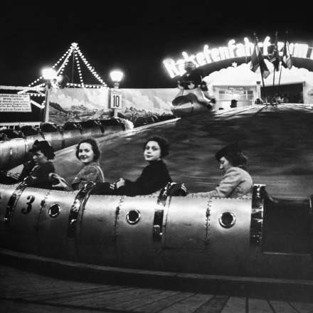 Schwarz-weiss Foto, auf dem vier Frauen zu sehen sind, die in einer Achterbahn sitzen. Hinter der Achterbahn steht über dem Kassenhäuschen ein Schriftzug: "Raketenfahrt zum Mond".
