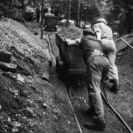 Das historische Foto in schwarz-weiß zeigt Bergleute, die um 1936 mit reiner Muskelkraft gefüllte Loren aus einer Grube bewegen.