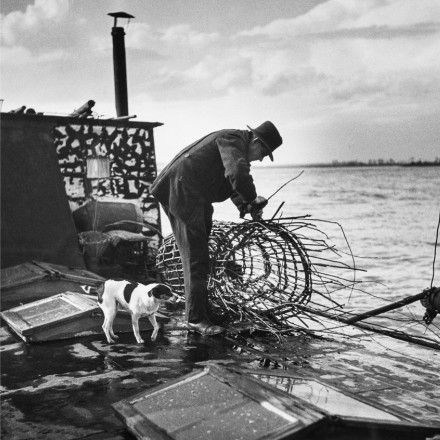 Schwarz-weiss Foto, auf dem ein Fischer zu sehen ist, der einen Korb am Wasser flechtet. Neben ihm steht ein kleiner Hund. 