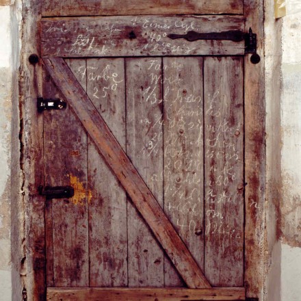 Eine alte Holztür aus groben senkrechten Brettern. Die Tür zeigt deutliche Gebrauchsspuren und Risse im Holz. Auf der Tür sind handgeschriebene kaum noch erkennbare Kreidenotizen erkennbar. Es handelt sich dabei um ein altes Rezept, um die Farbe Grün zu mischen. Oben auf der Tür liegt ein kleines rotes Heft.