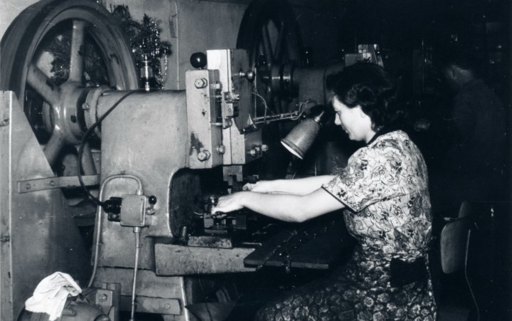 Frau in gemustertem Kleid arbeitet an einer großen industriellen Scheren-Schneidemaschine in einer Werkstatt.