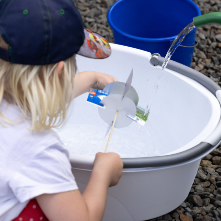 Kind mit Baseballkappe hält selbstgebasteltes Papierrad über mit Wasser gefülltes Becken.