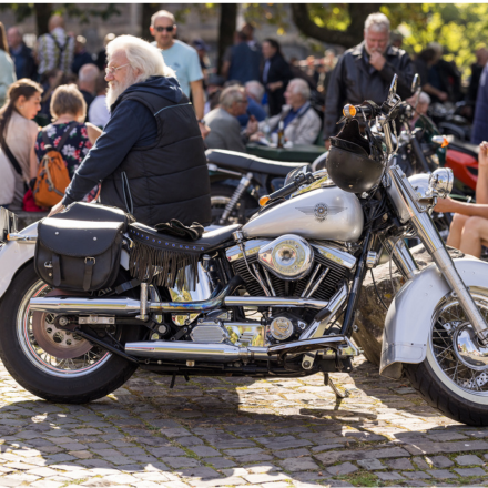 Mann sitzt auf einem silbernen Motorrad, im Hintergrund sind unterschiedliche Menschen zu sehen