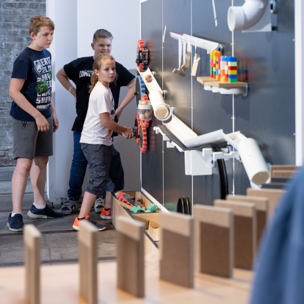 Drei Kinder stehen vor einer Wand mit einer Kugelbahn aus Rohren und bunten Bausteinen, im Vordergrund eine Reihe Dominosteine.