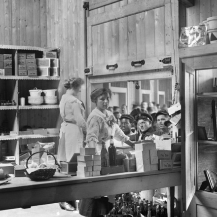 Innenraum einer Kantine mit Holzwänden, Regalen voller Vorratsdosen und Flaschen, einem großen Tisch in der Mitte und mehreren Personen, darunter zwei Frauen in Schürzen.