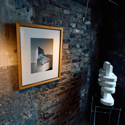 Gerahmtes Bild mit abstrakter Architektur an einer Backsteinwand neben einer weißen abstrakten Skulptur auf einem Metallgestell.