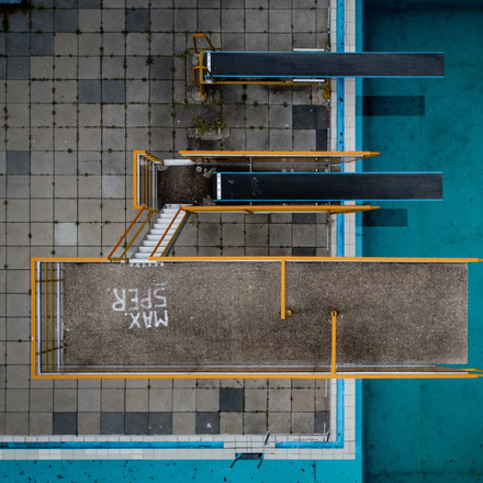Luftaufnahme eines Schwimmbeckens mit Sprungturmanlage. Drei lange Sprungbretter ragen von einer gefliesten Plattform über das Wasser.