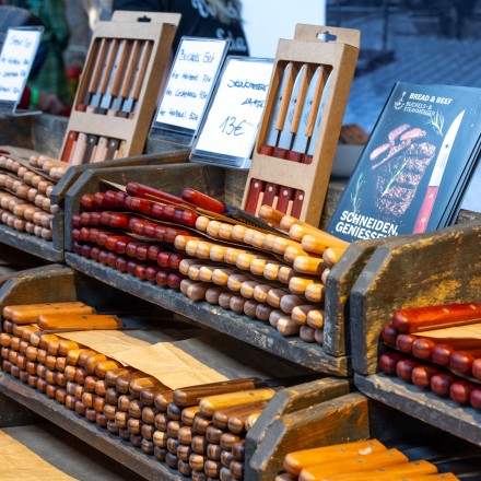 Ein Marktstand mit einer Auswahl handgefertigter Messer. Die Messer haben unterschiedlich gefärbte Holzgriffe und liegen in Holzkästen aus. Im Hintergrund sieht man Preisschilder und ein Buch.