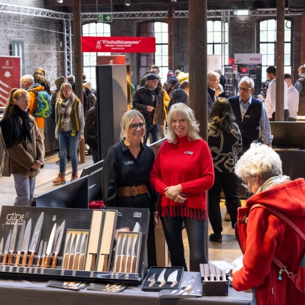 Menschen besuchen eine Messe in einer Industriehalle und informieren sich an verschiedenen Ständen über Produkte. Im Vordergrund präsentieren zwei Ausstellerinnen Messerprodukte. 
