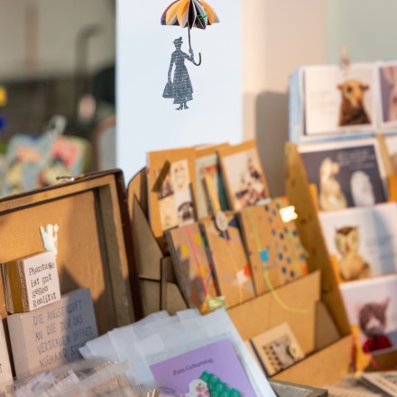 Ein kleiner Verkaufsstand mit handgemachten Papeterieartikeln: In Holzkisten und Aufstellern sind Postkarten, kleine Geschenkkarten und Drucke arrangiert. Im Vordergrund liegt eine offene Holzbox mit verpackten Karten und bunten Motiven. Darüber hängt ein filigranes Mobile aus Papier, das eine stilisierte Figur mit Regenschirm zeigt.