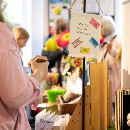 Ein Marktstand mit handgefertigten Papierprodukten. Im Vordergrund hängt ein Schild aus Papier an einem Ständer mit der Aufschrift "Nimm dir, was du brauchst!". Drumherum stehen die Wörter "Kuss", "Zeit", "Glück" und "Sonne". Im Hintergrund sind Personen und weitere Dinge aus Papier unscharf zu sehen.