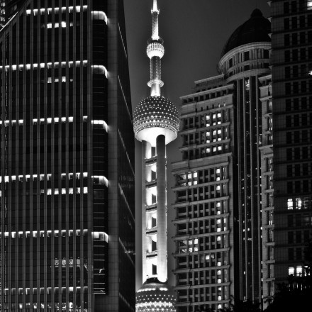 Schwarzweißaufnahme von einer Skyline mit dicht stehenden Hochhäusern. In der Bildmitte ist ein beleuchteter Fernsehturm zu sehen. Er ist umgeben von beleuchteten Hochhäusern.