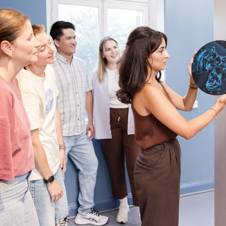 Eine Gruppe junger Erwachsener, die um eine andere junge Frau steht. Diese hält eine runde Scheibe in der Hand, auf der Wölfe in schwarz blau sind.