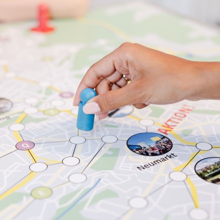 Foto, auf dem man eine Hand sieht, die eine blaue Spielfigur auf einem Spielbrett bewegt. Das Spielbrett ist ein Netz aus verbundenen Punkten und Fotos.