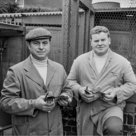 Zwei Männer in Arbeitskleidung halten Tauben vor einem Taubenschlag mit Maschendrahtzaun und Backsteinmauer.